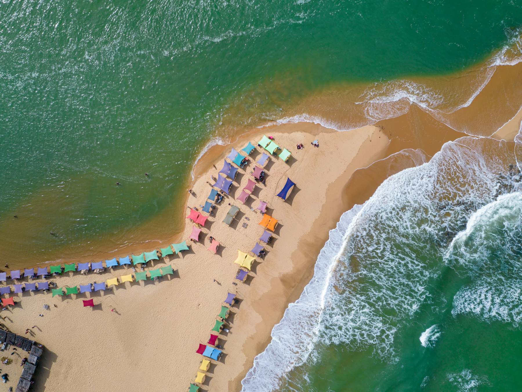 Vista aérea da praia de Caraíva, com guarda-sóis coloridos na faixa de areia entre o rio e o mar verde e as ondas quebrando na beira.
