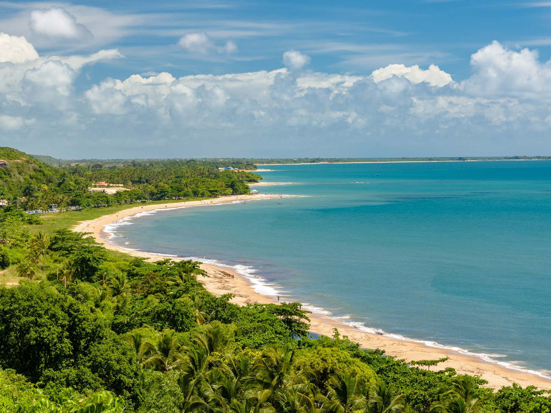 Vista do alto da Cidade Histórica de Porto Seguro para o litoral, com a mata verde, a faixa de areia em curva e o mar azul-turquesa.