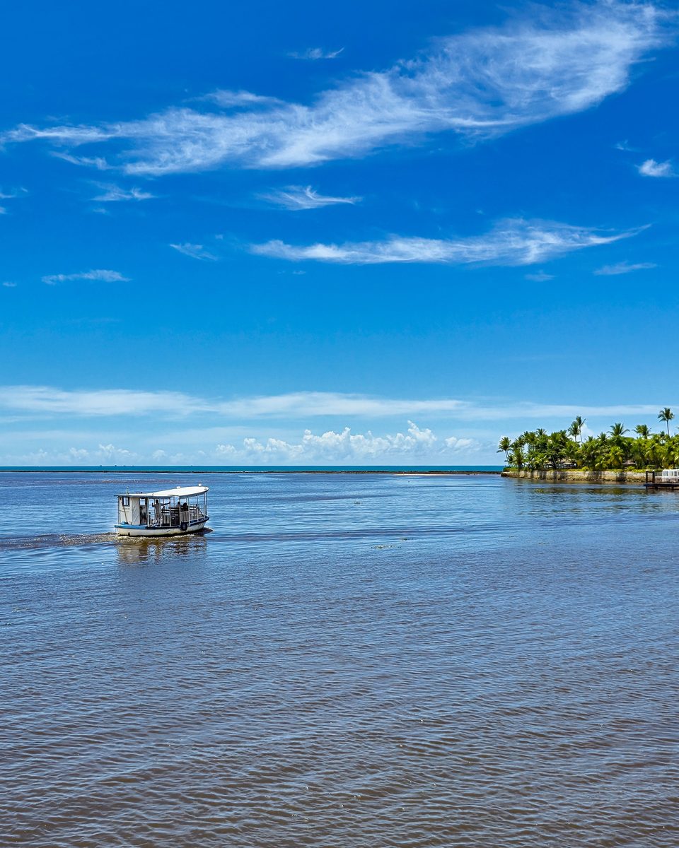 Barco de passeio navegando no mar calmo e azul de Porto Seguro, com o coqueiral e as casas da margem ao fundo e o céu com nuvens finas.