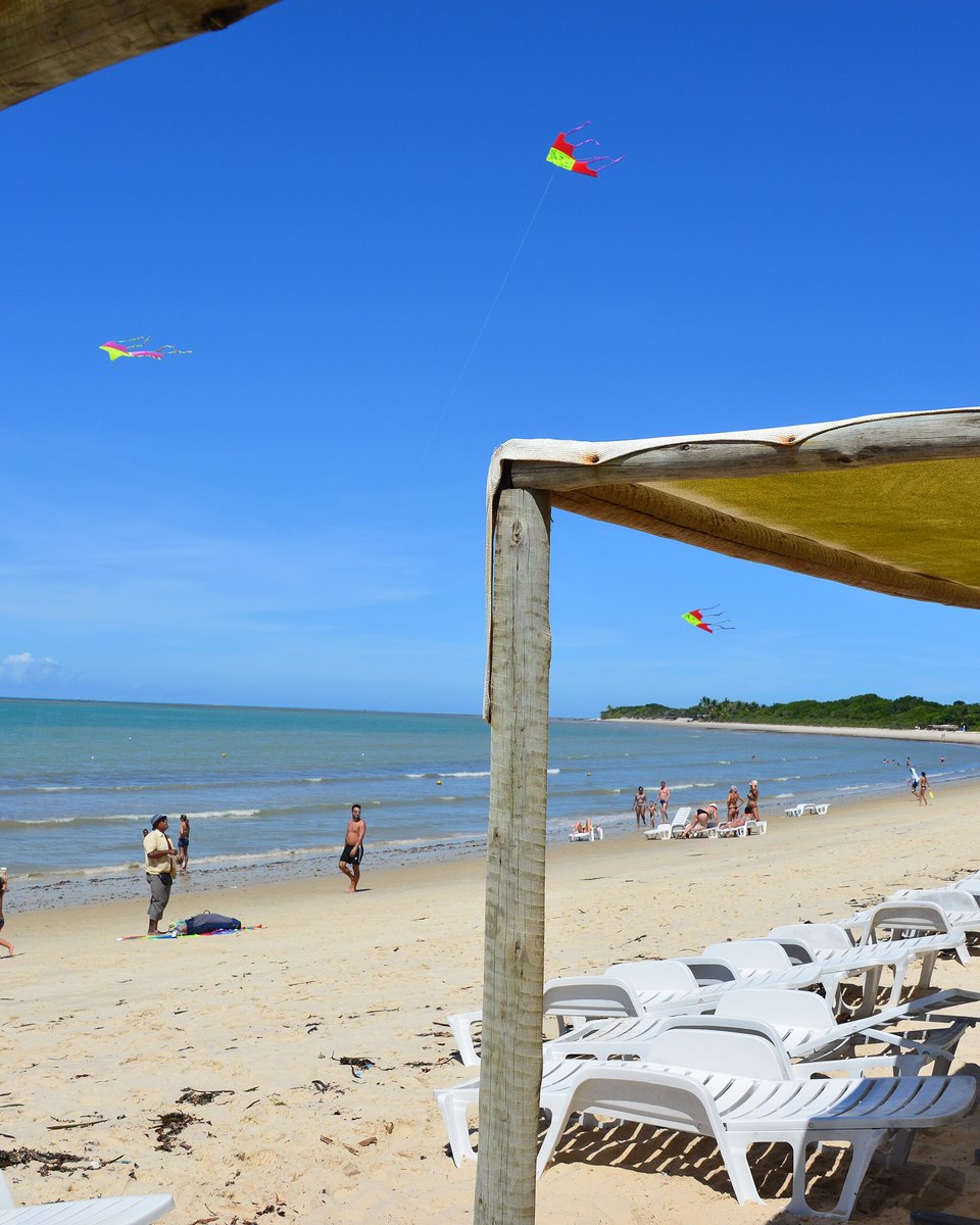 Praia de Porto Seguro com barracas, espreguiçadeiras e pipas no céu azul.