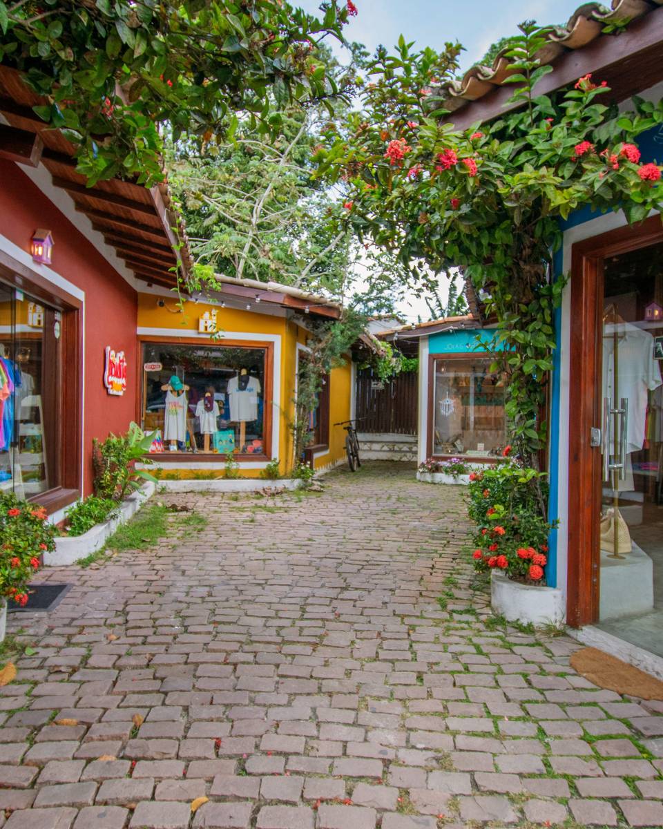 Pátio de pedra no centro de Arraial d'Ajuda com lojinhas de fachada vermelha, amarela e azul, vitrines de roupa e moda praia e floreiras com flores vermelhas.