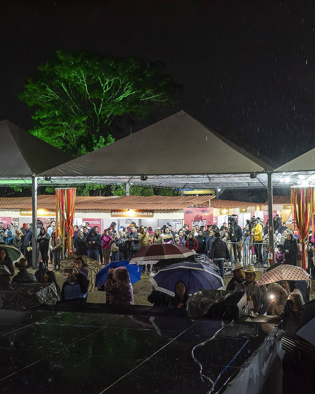 Tendas iluminadas de uma feira cultural à noite em Presidente Prudente, com o público de guarda-chuva circulando entre as barracas na chuva.