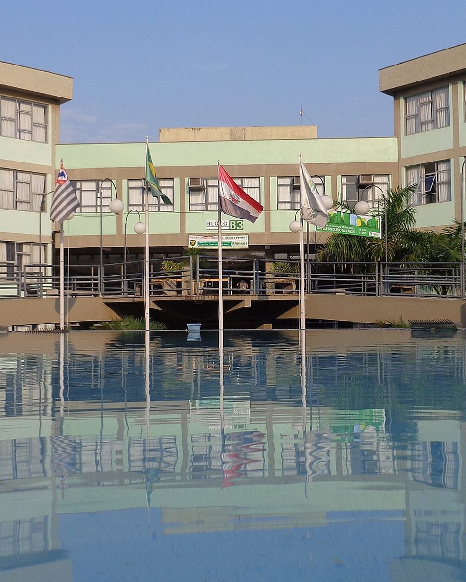 Prédio de três andares do Campus II da Unoeste, em Presidente Prudente, refletido num espelho d'água, com bandeiras hasteadas na frente.