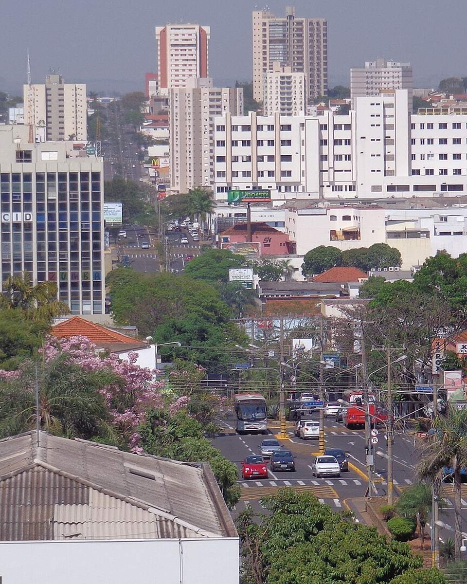 Vista do alto de Presidente Prudente, com uma avenida movimentada de carros e ônibus entre árvores, prédios comerciais e residenciais ao fundo.