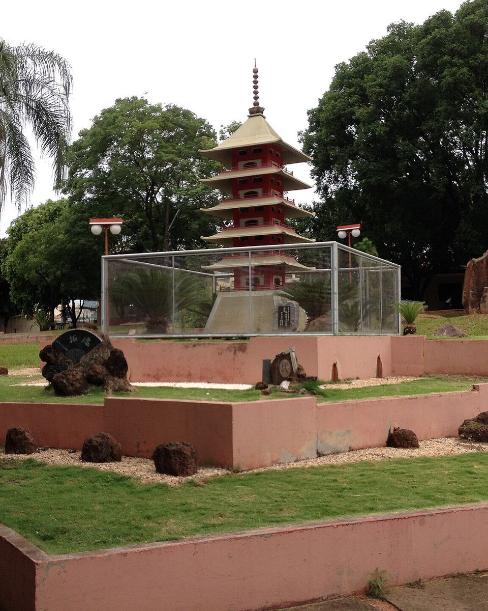 Pagode vermelho japonês na Praça das Cerejeiras, em Presidente Prudente, entre palmeiras e jardim.