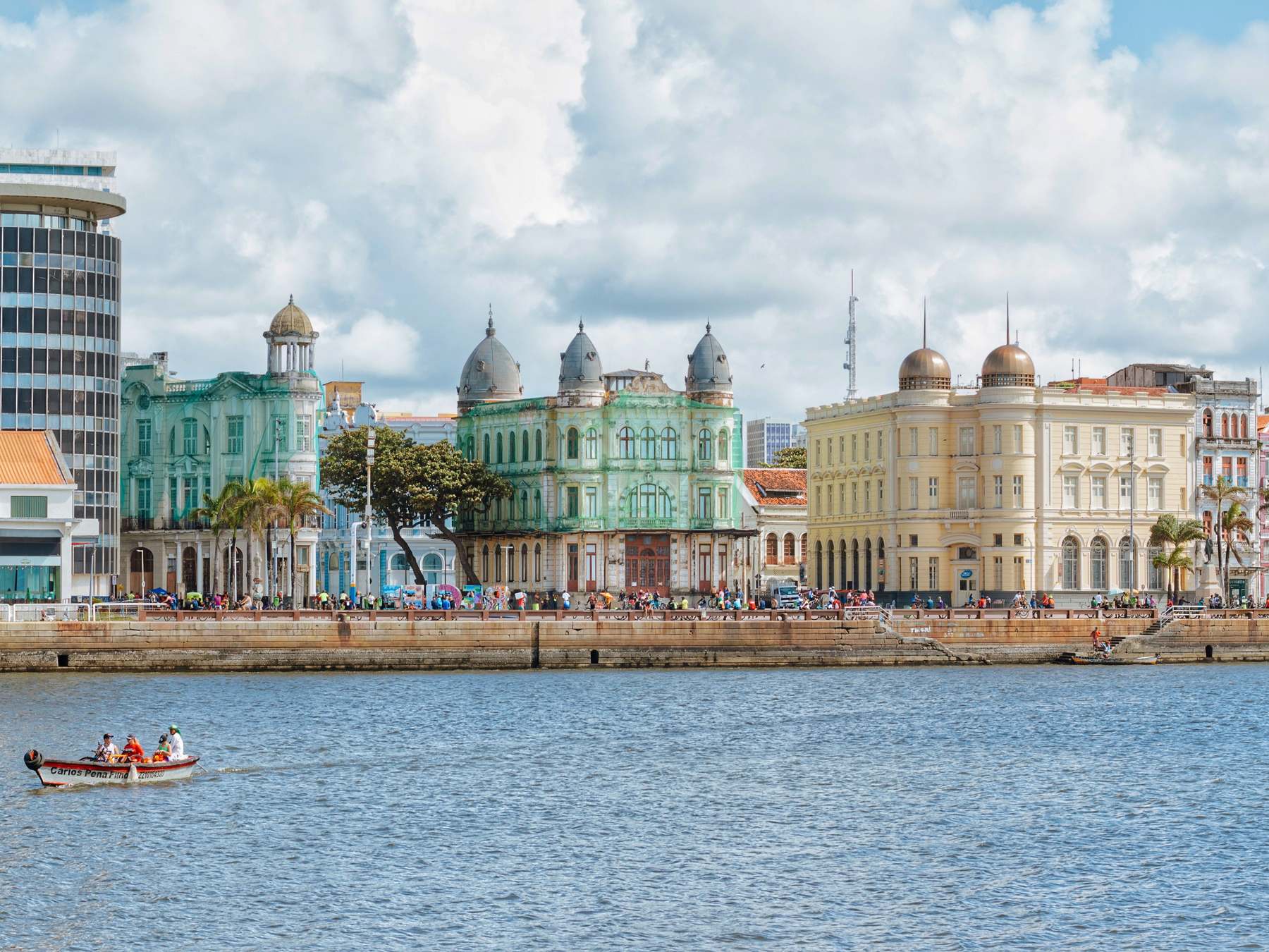 Casarões do Marco Zero, com cúpulas e fachadas verde, amarela e branca, vistos das águas do porto do Recife, com um barco pequeno passando em primeiro plano e o céu azul com nuvens brancas.