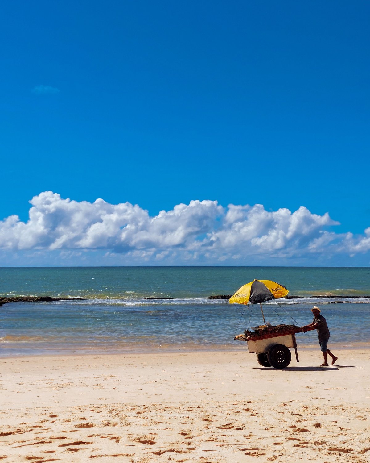 Vendedor ambulante empurrando um carrinho com guarda-sol amarelo pela areia da Praia de Boa Viagem, no Recife, com o mar verde e o céu azul com nuvens brancas.