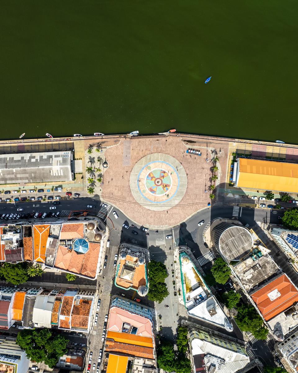 Vista de cima da Praça do Marco Zero e do Bairro do Recife, com a rosa dos ventos no piso da praça, os armazéns do cais à beira da água escura do porto e os telhados laranja dos casarões.