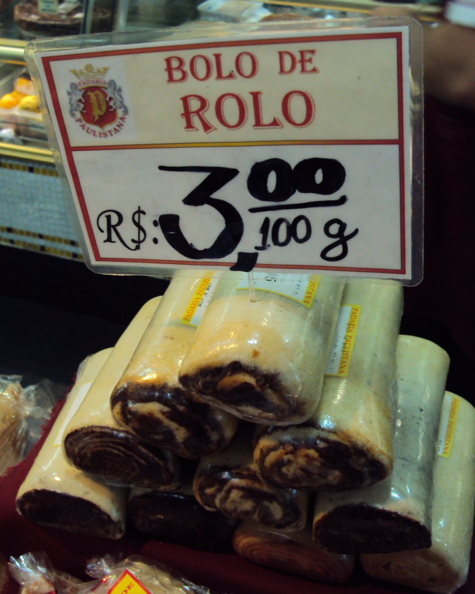 Fatias de bolo de rolo embaladas em plástico numa vitrine, com camadas finas enroladas em espiral, e uma plaquinha com o preço de R$ 3,00 por 100 gramas escrita à mão.