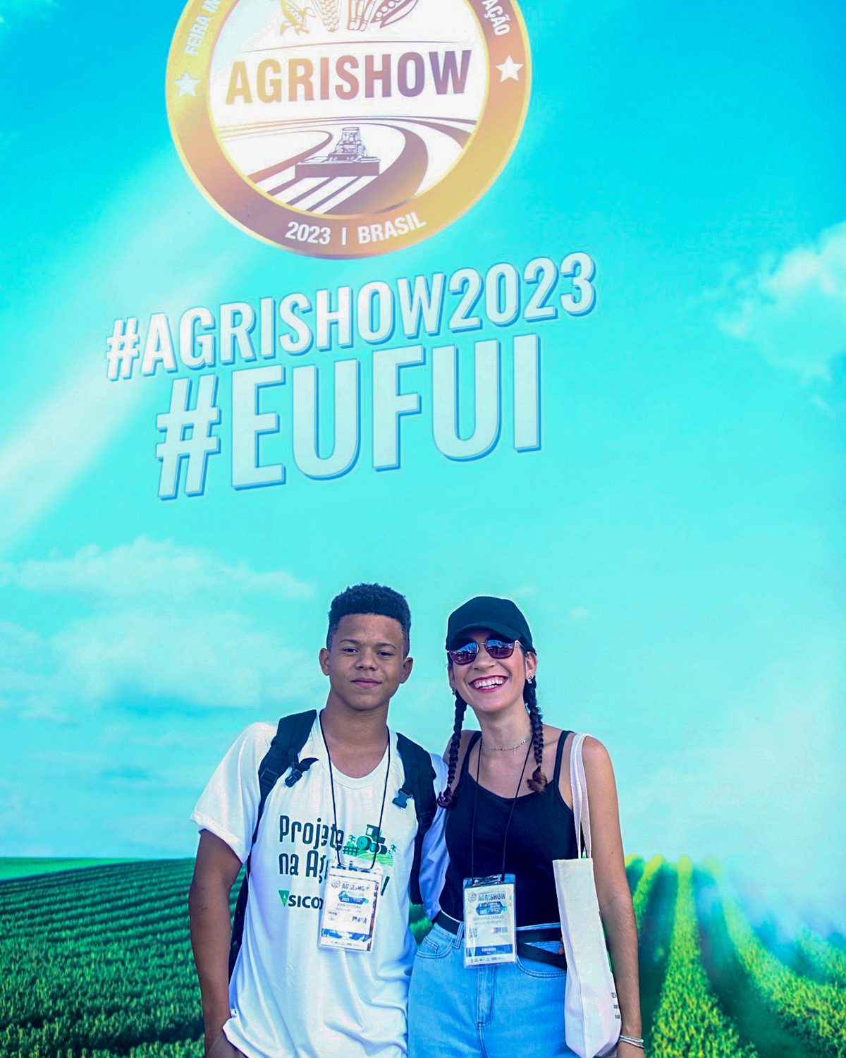 Rapaz e moça sorrindo lado a lado com crachá no pescoço, diante de um painel azul da Agrishow com a hashtag Eu Fui, em Ribeirão Preto.