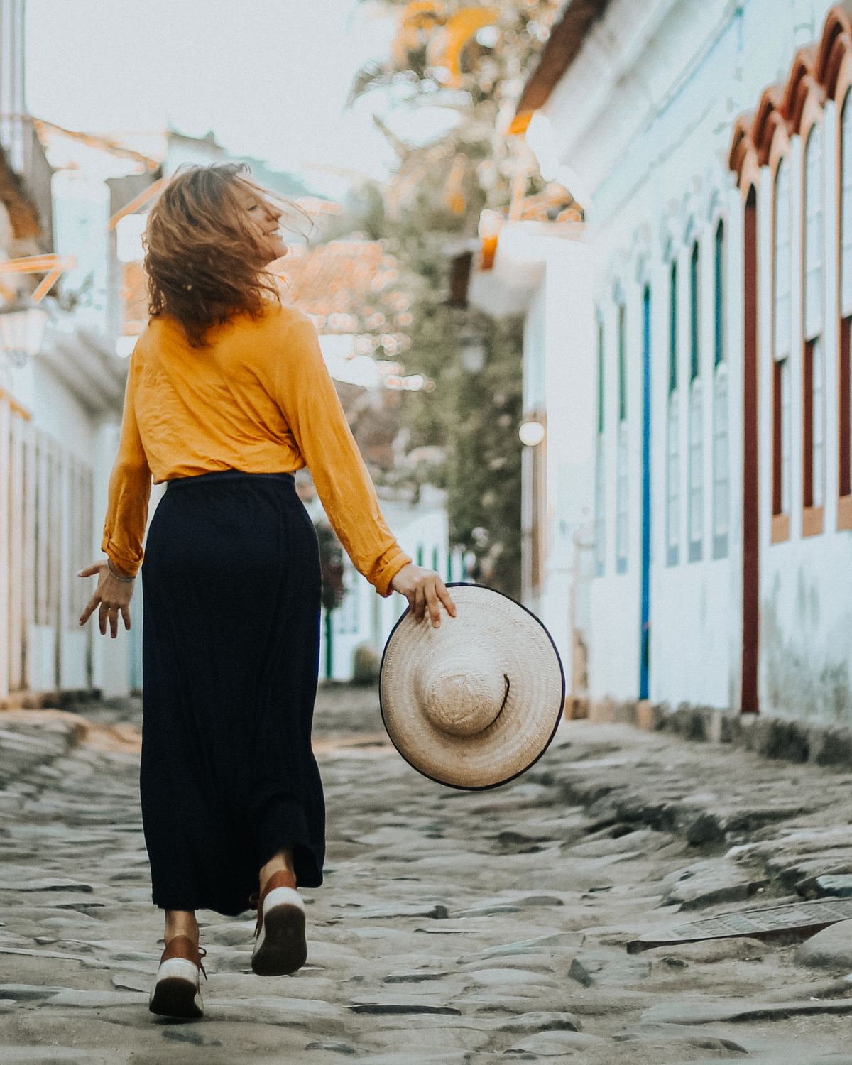 Mulher de blusa amarela e saia preta caminhando de costas por uma rua de pedras do centro histórico de Paraty, com um chapéu de palha na mão.