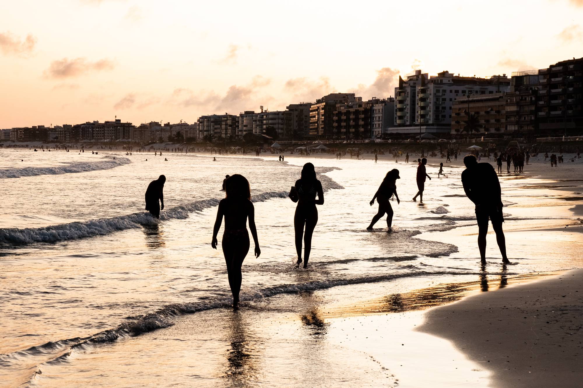 Silhuetas de pessoas caminhando na beira do mar ao pôr do sol na Praia do Forte, em Cabo Frio, com os prédios da orla ao fundo.