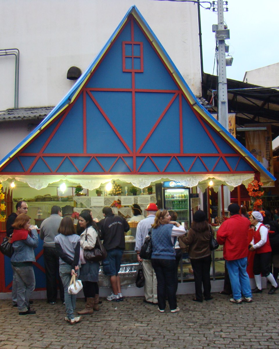 Pessoas fazendo fila e passeando diante de uma barraca em forma de chalé azul e vermelho, na Bauernfest, festa de tradição alemã em Petrópolis.
