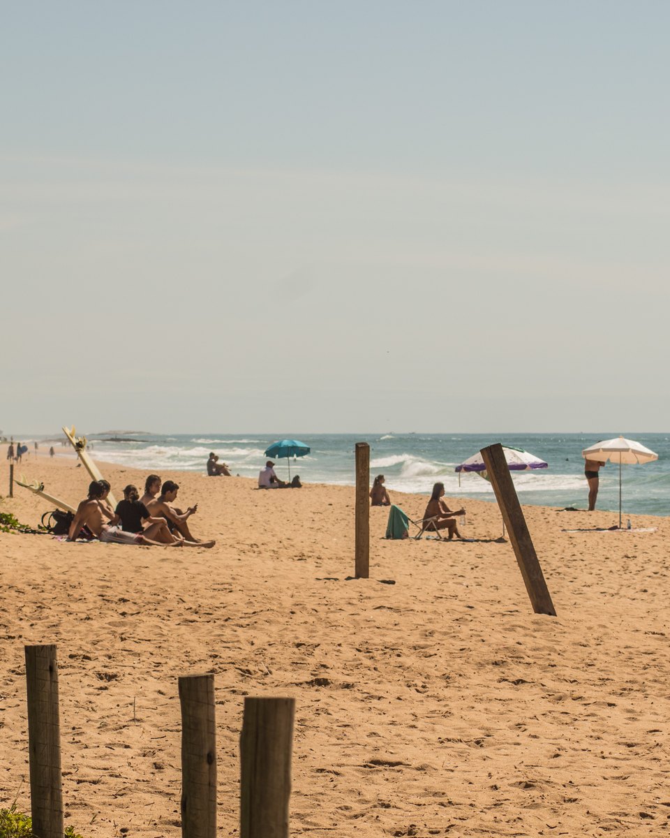 Pessoas sentadas na areia e guarda-sóis na Praia do Pecado, em Macaé, com prédios à esquerda e um navio no horizonte.