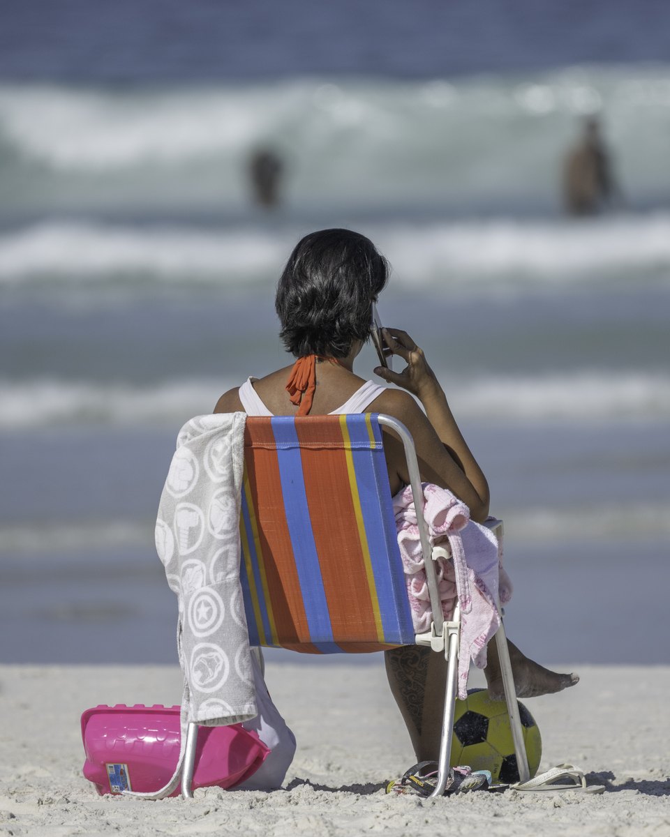Mulher de costas numa cadeira de praia listrada, falando ao celular na areia da Praia do Forte, em Cabo Frio, com banhistas no mar ao fundo.