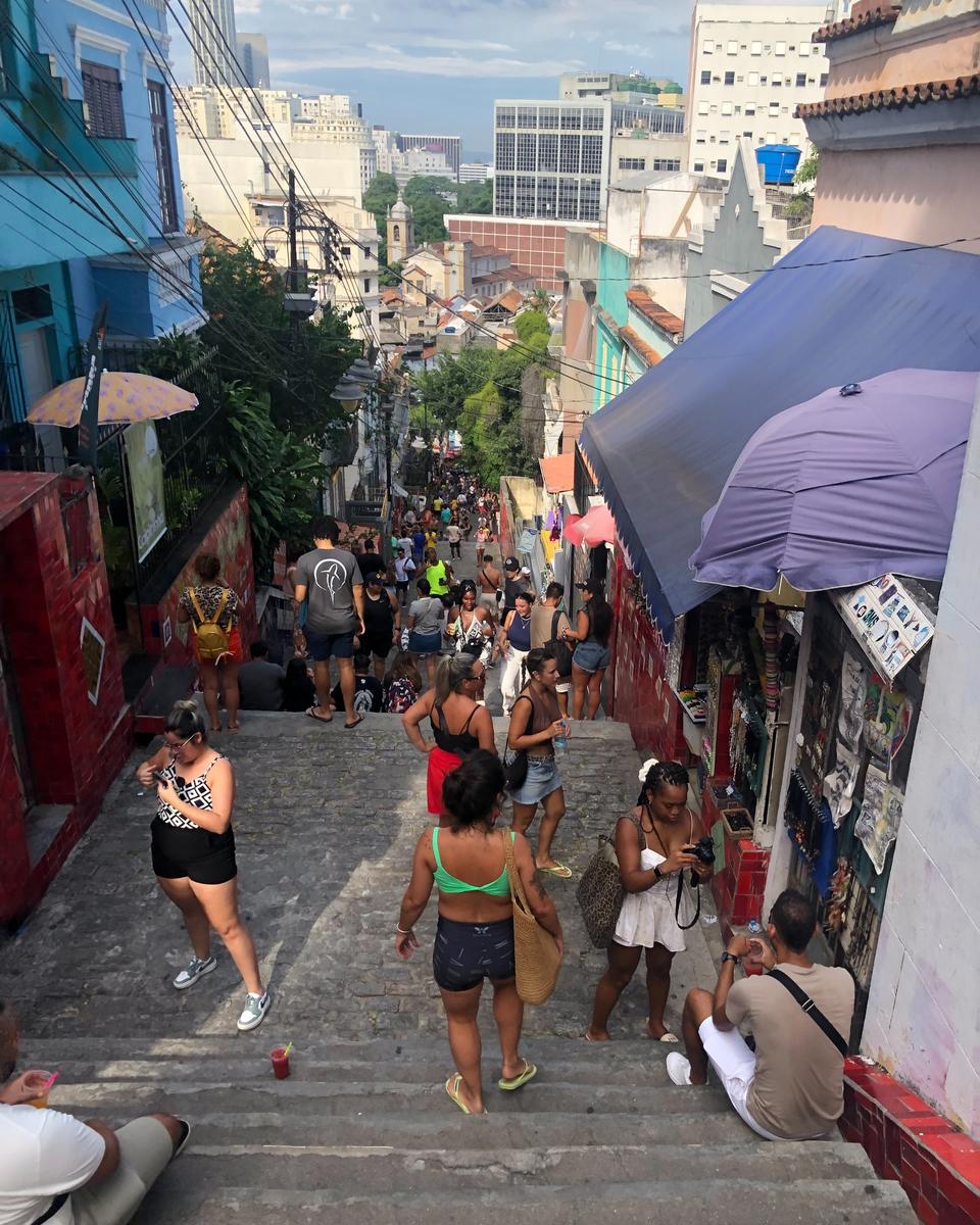 Visitantes subindo e descendo a Escadaria Selarón, na Lapa, no Rio de Janeiro, alguns fotografando com o celular, entre degraus de azulejos vermelhos, barracas de lembrancinhas e casas coloridas.