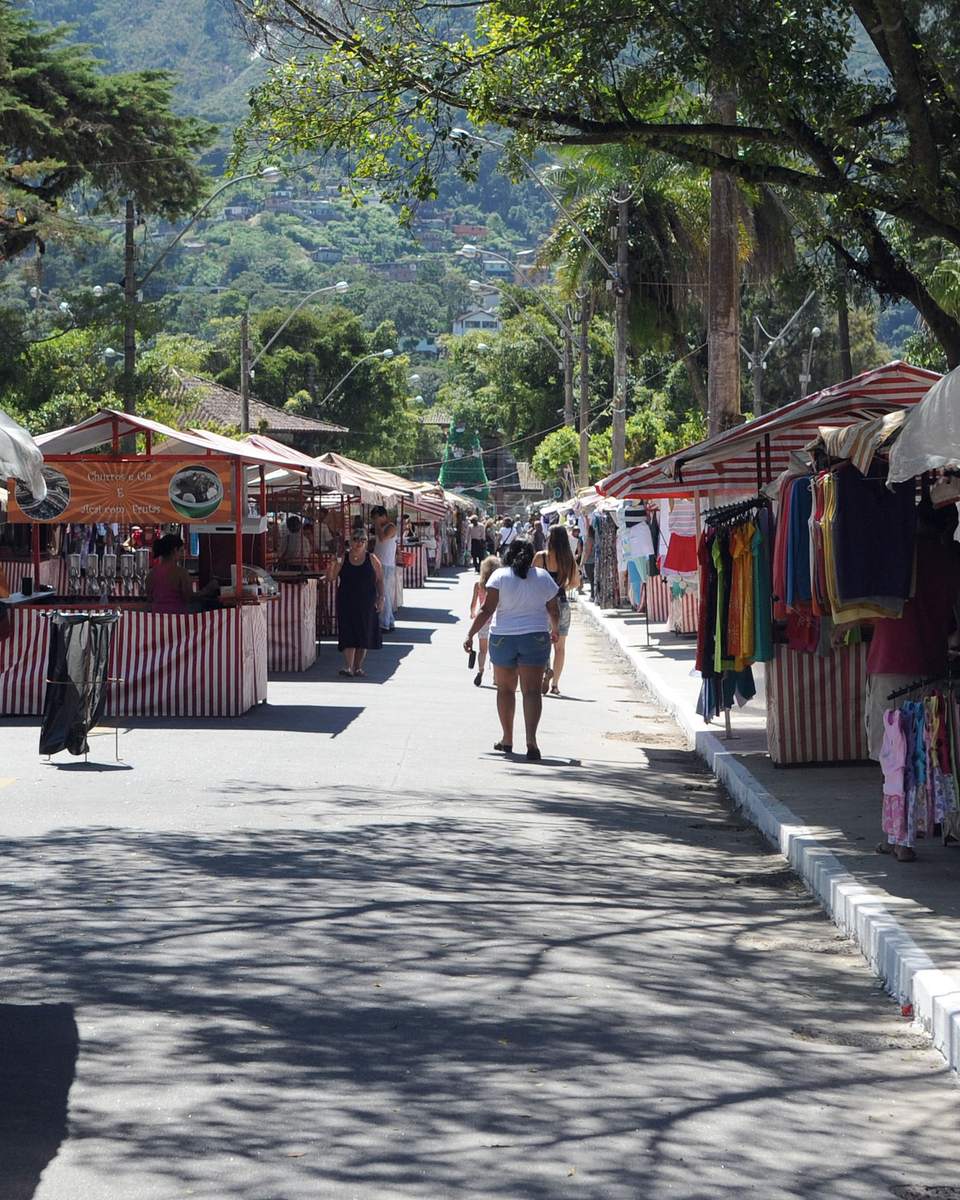 Rua arborizada com barracas listradas de roupas, móveis e artesanato na Feirinha do Alto, em Teresópolis, com pessoas caminhando entre elas e as montanhas da serra ao fundo.
