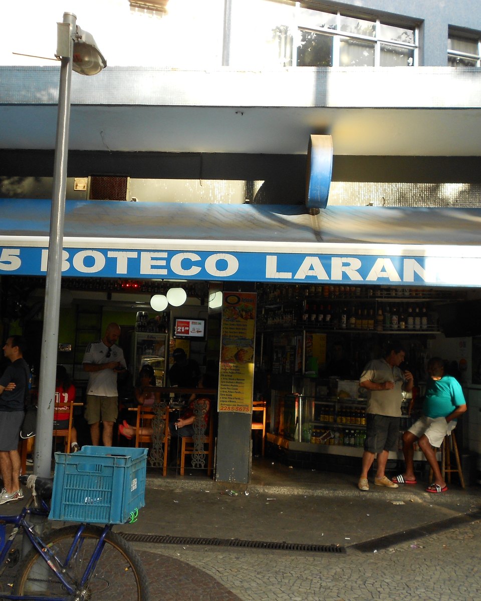 Fachada de um boteco de esquina em Laranjeiras, no Rio, com toldo azul, gente em pé no balcão, banquetas na calçada e uma bicicleta estacionada na frente.
