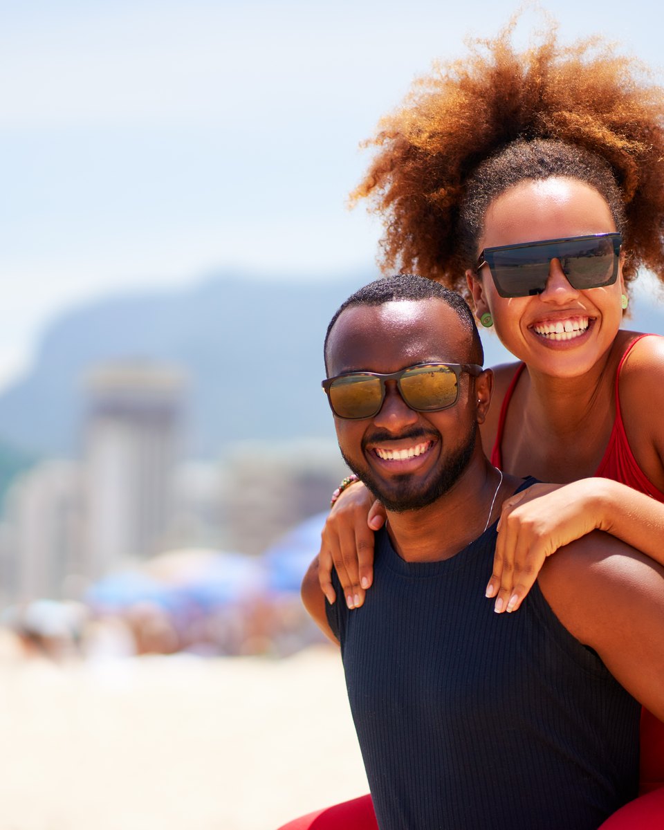 Casal sorrindo na praia de Ipanema, ela de cabelo crespo e vestido vermelho nas costas dele, os dois de óculos escuros, com prédios e morros desfocados ao fundo.