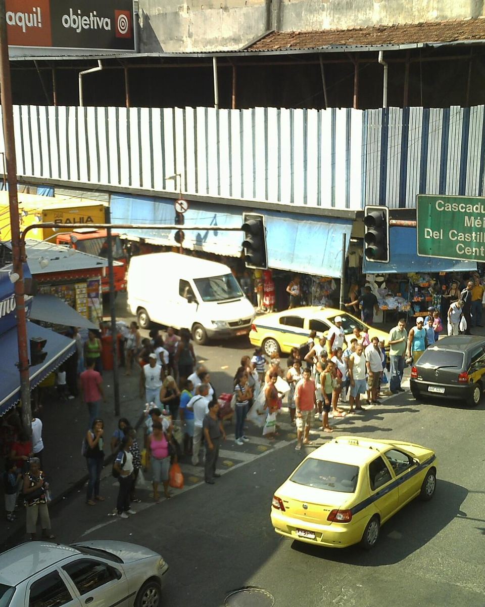 Vista do alto de uma esquina movimentada em Madureira, na Zona Norte do Rio, com gente atravessando a rua entre lojas e barracas, táxis amarelos e uma placa indicando Cascadura, Méier, Del Castilho e Centro.
