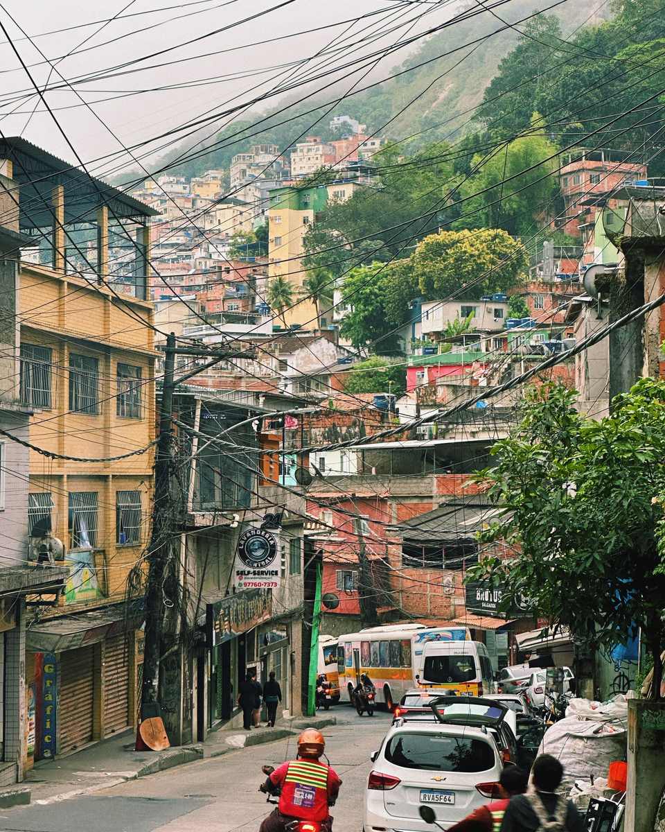 Rua movimentada numa favela do Rio de Janeiro, com mototaxista de colete, carros, van e ônibus subindo a ladeira, lojas no térreo, fios cruzando o céu e casas no morro ao fundo.
