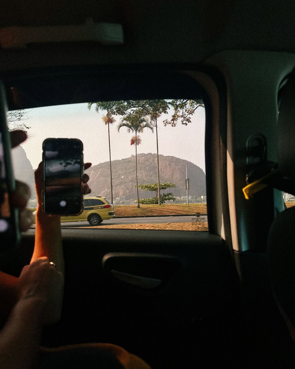 Mão segurando o celular dentro de um carro, filmando pela janela um táxi amarelo, palmeiras e o Morro da Urca, no Rio de Janeiro.