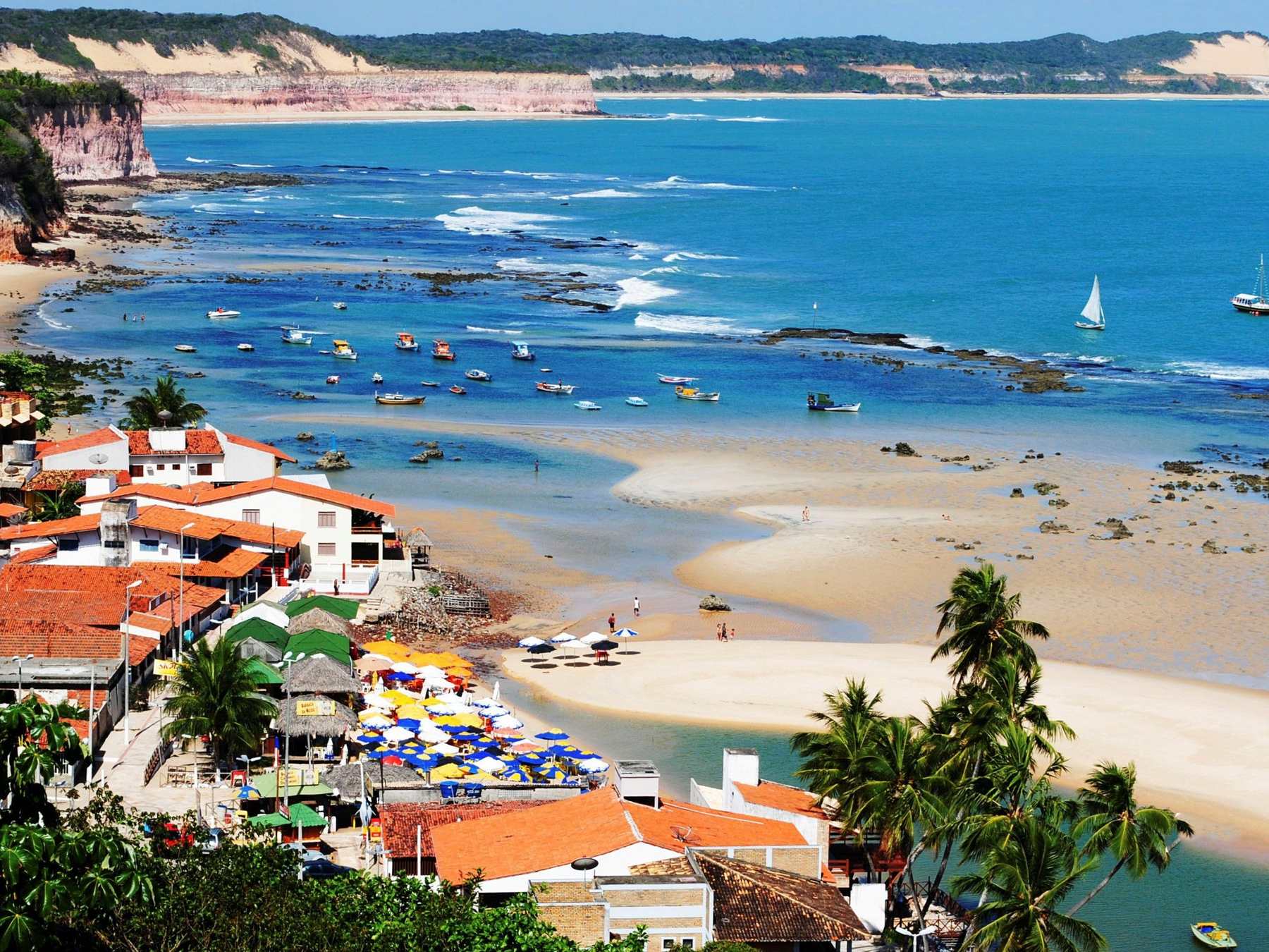 Vila de Pipa vista do alto, com casas de telhado laranja, jangadas ancoradas no mar azul e falésias coloridas ao fundo