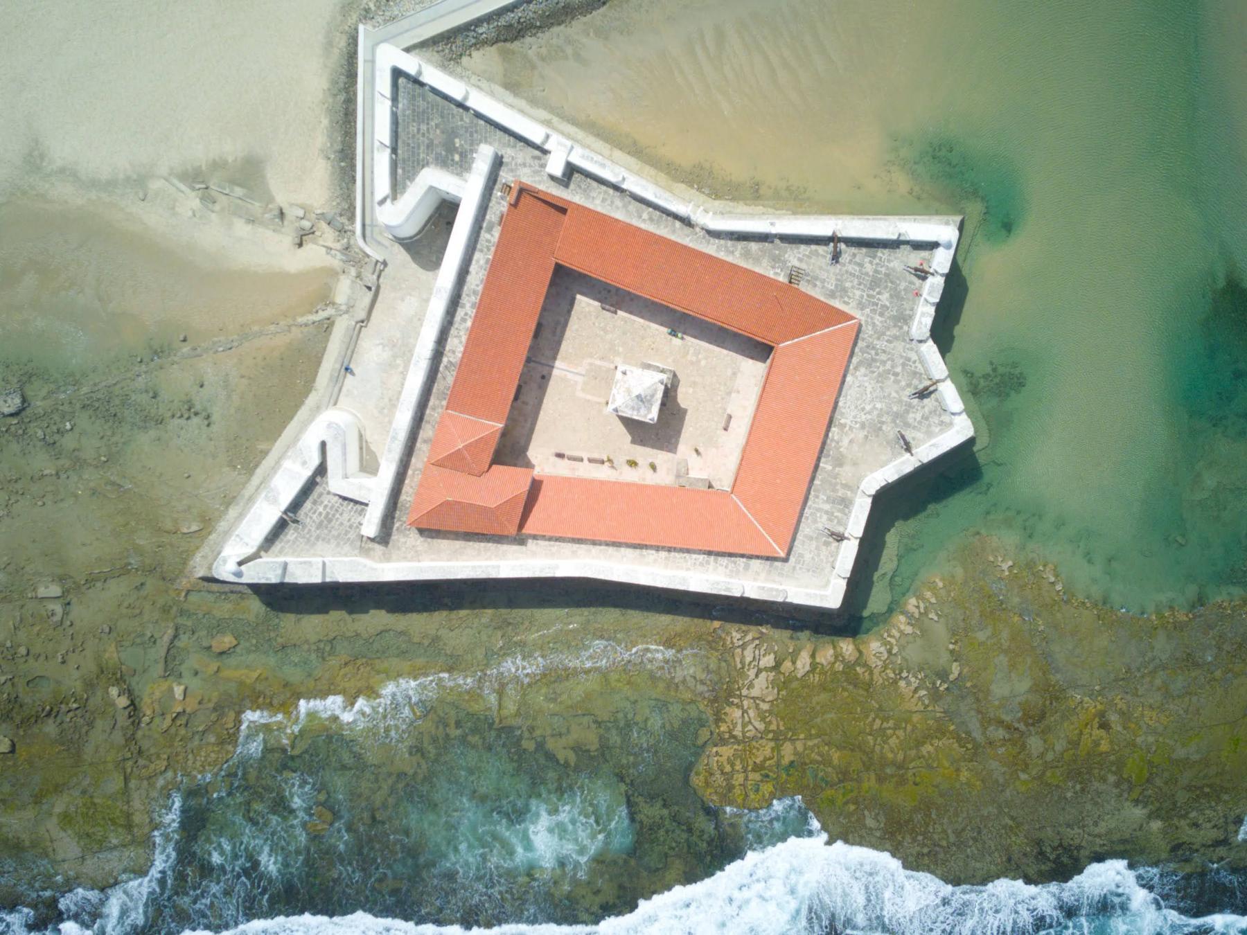 Foto aérea vertical do Forte dos Reis Magos, fortaleza em forma de estrela com telhado laranja cercada por recifes e ondas na foz do rio Potengi