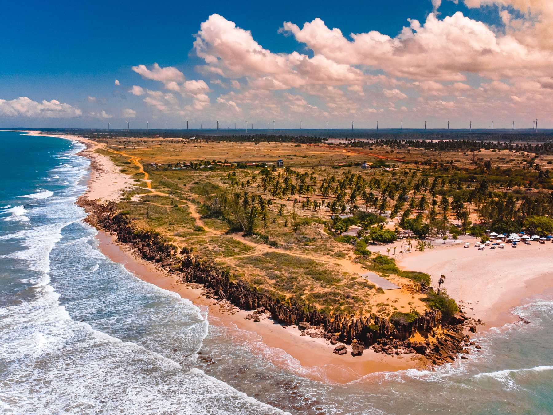 Vista aérea da praia de São Miguel do Gostoso, com faixa de areia, coqueiral, falésias baixas e aerogeradores na linha do horizonte