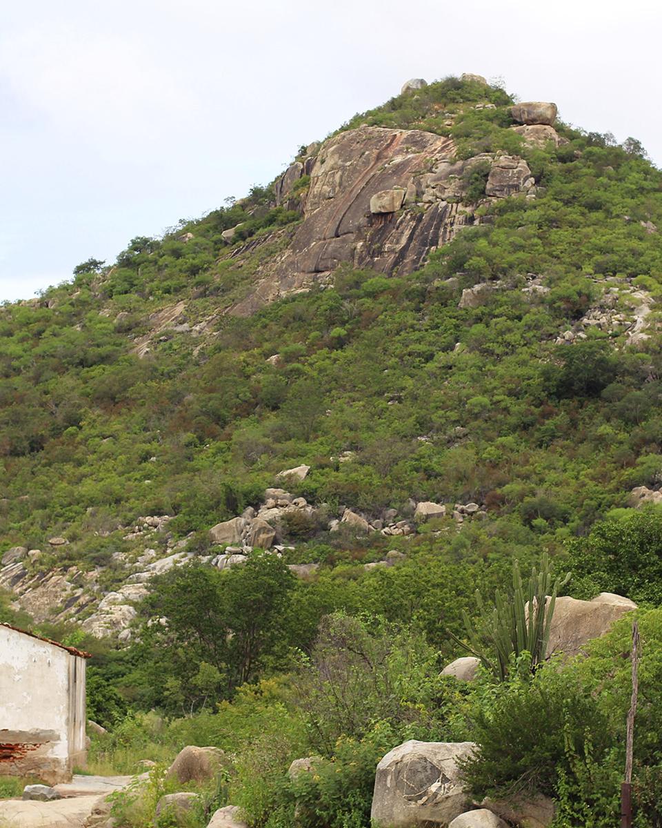 Serra de pedra coberta de caatinga no Seridó potiguar, com blocos de rocha entre a vegetação e uma casa de alvenaria no sopé