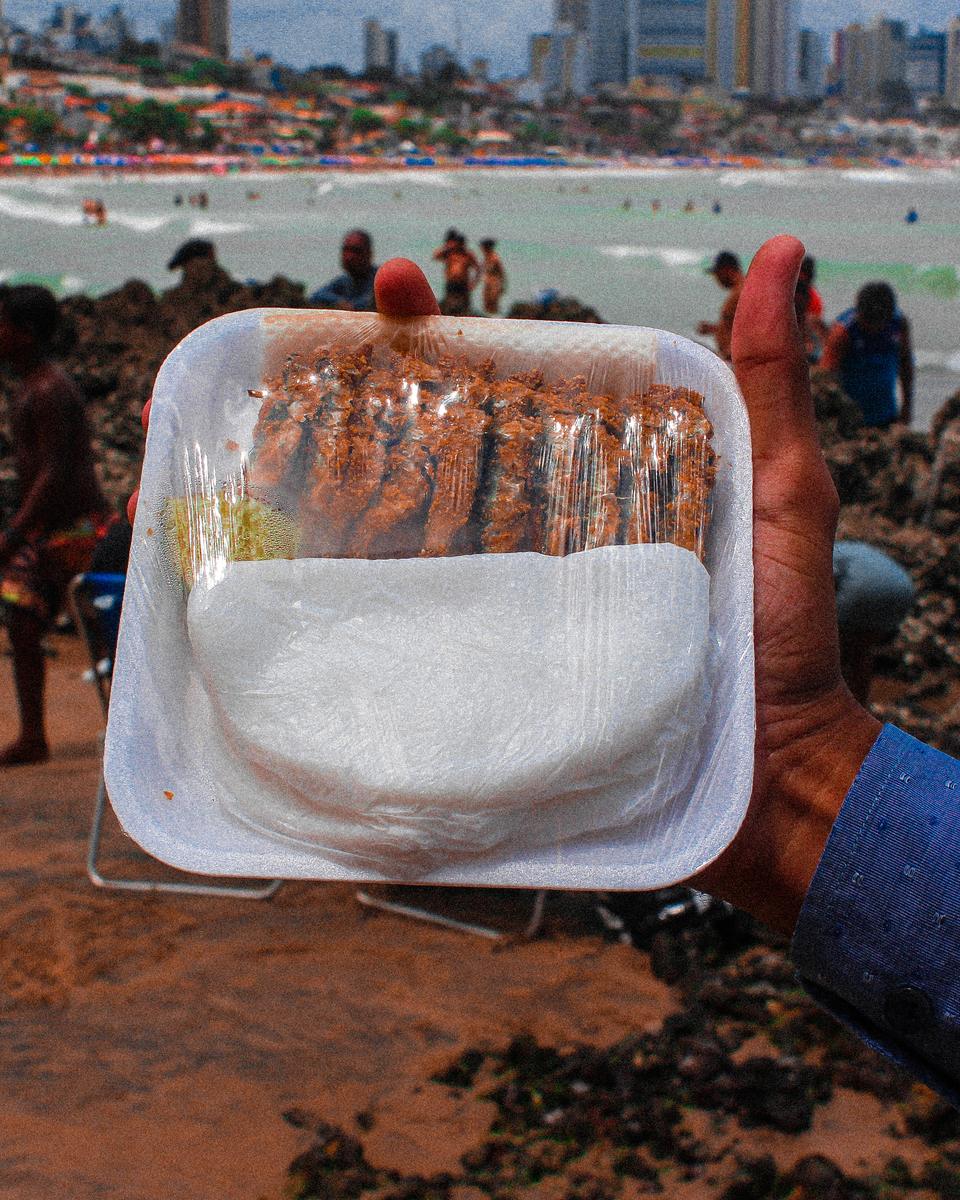 Mão segurando uma bandeja com tapioca e peixinhos fritos, a ginga com tapioca, na beira da praia, com a cidade ao fundo
