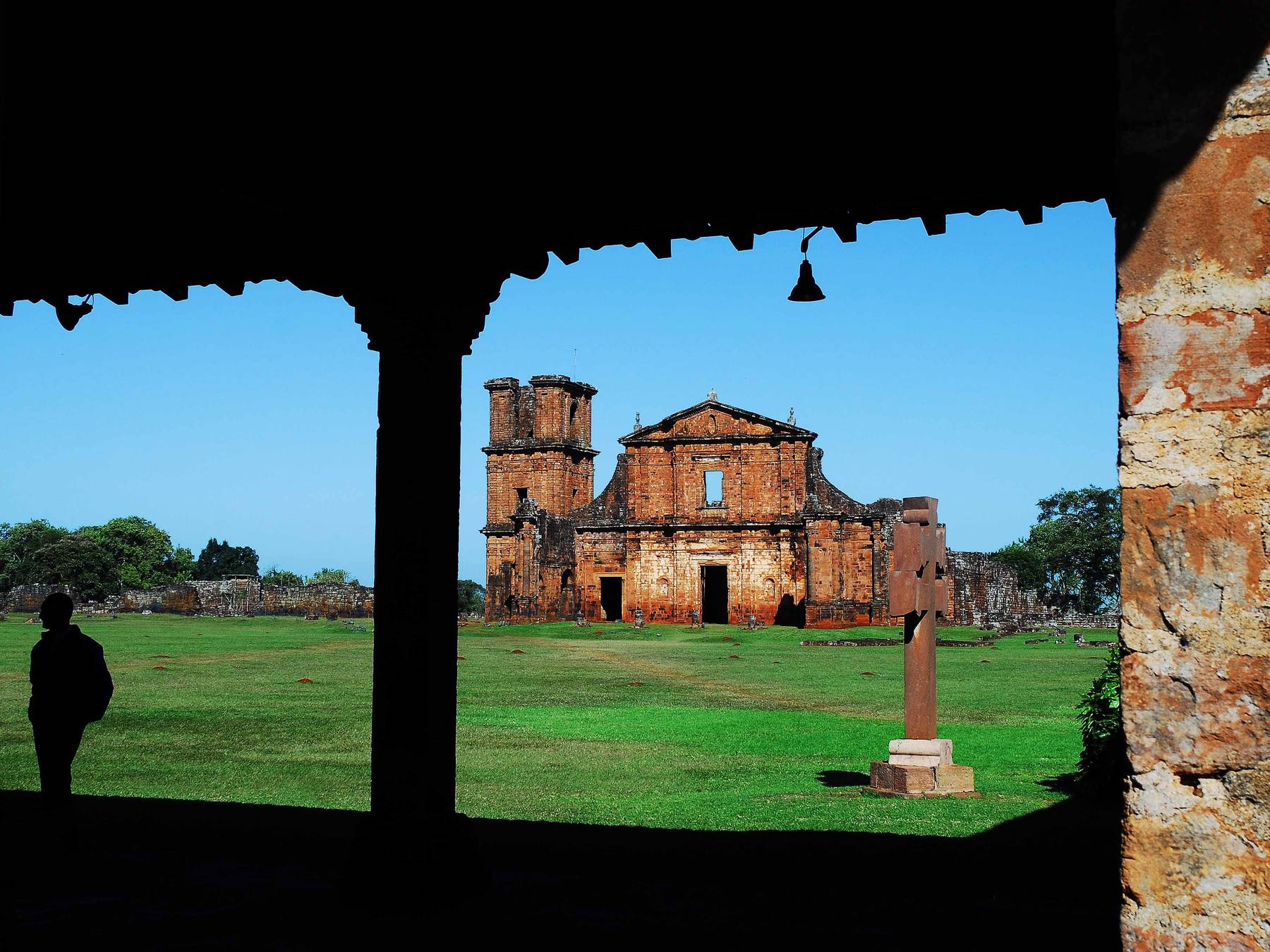 Ruínas da igreja de São Miguel Arcanjo vistas pela janela escura do museu das Missões, com o gramado verde, uma cruz e o céu azul, e a silhueta de um visitante à esquerda.