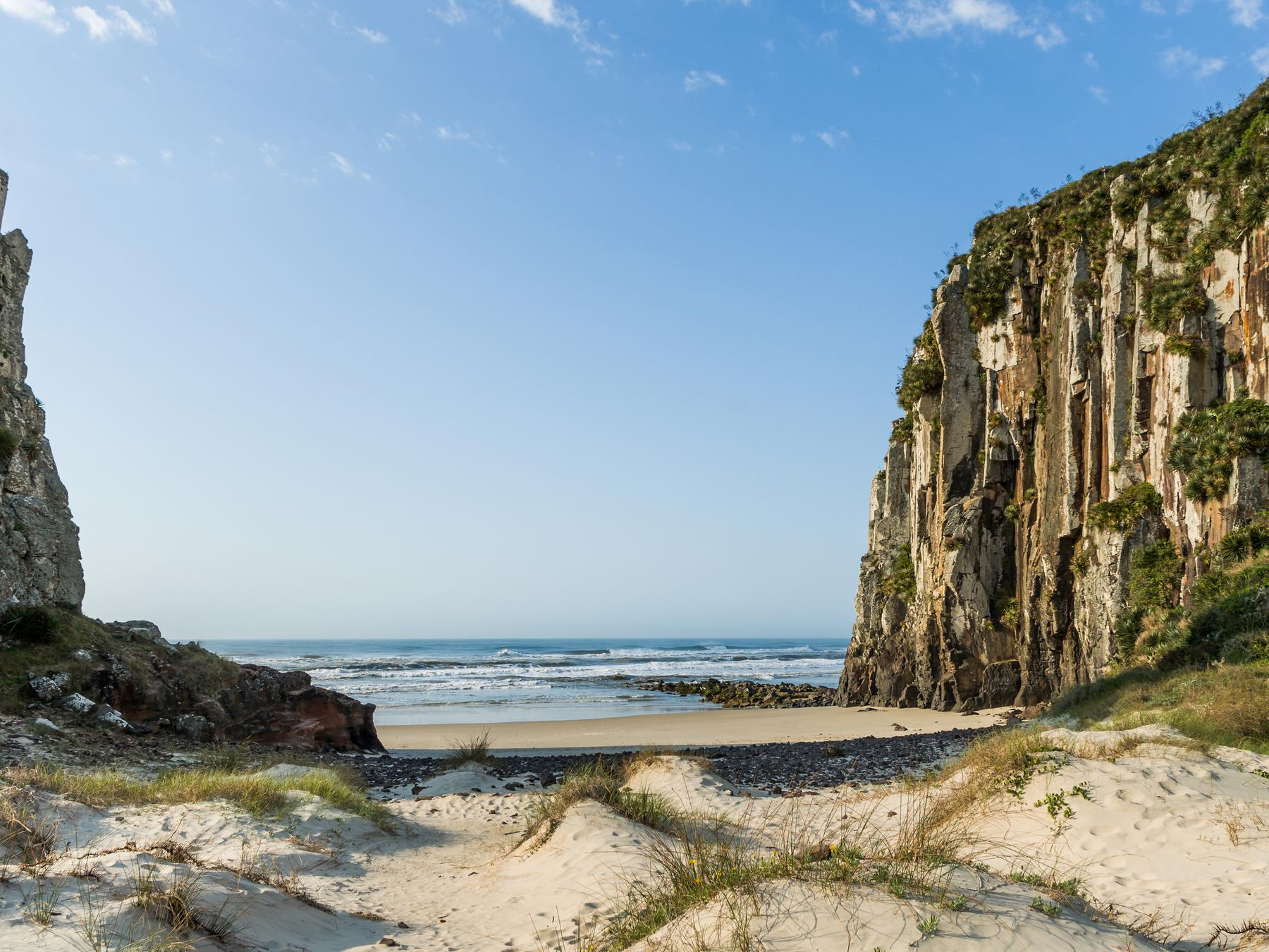 Praia da Guarita, em Torres, com a faixa de areia e as dunas entre dois paredões de rocha vulcânica e o mar ao fundo, num dia de céu azul.