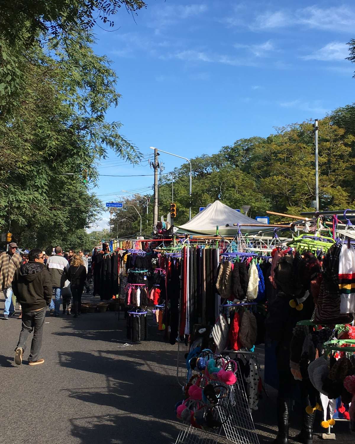 Barracas de roupa e artesanato do Brique da Redenção num domingo de sol, com gente caminhando pela rua arborizada ao lado do Parque Farroupilha, em Porto Alegre.