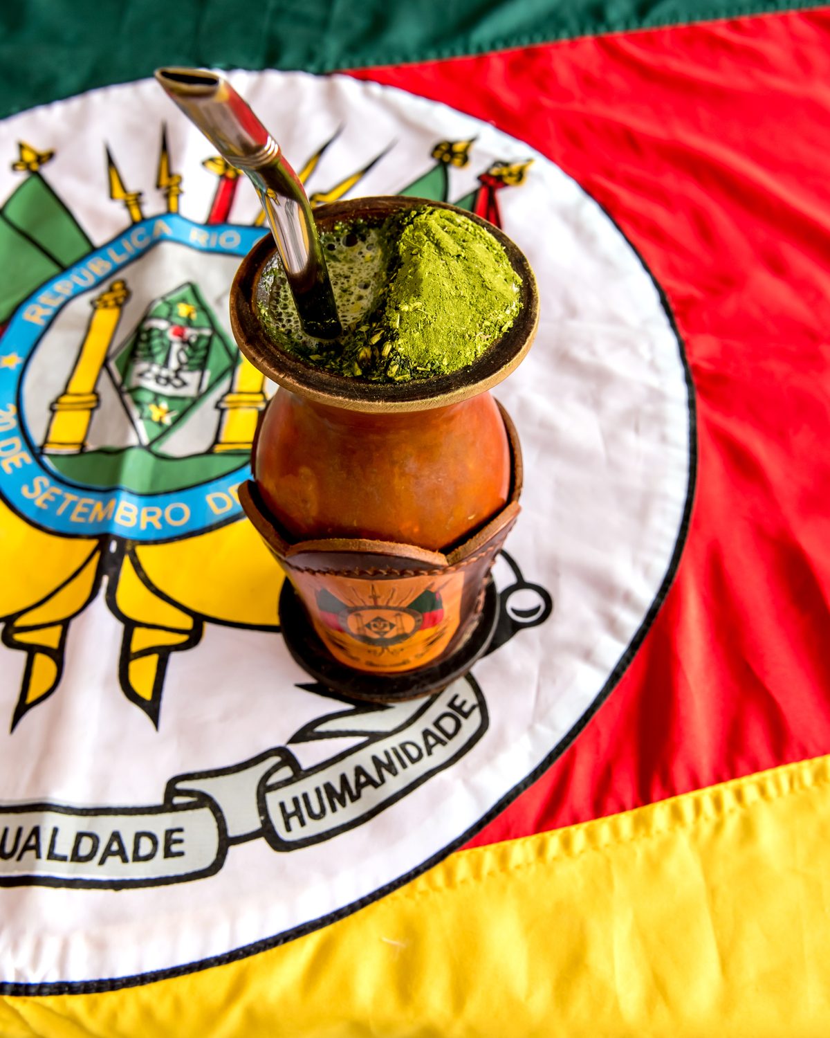 Cuia de chimarrão com erva-mate e bomba de metal apoiada sobre a bandeira do Rio Grande do Sul.
