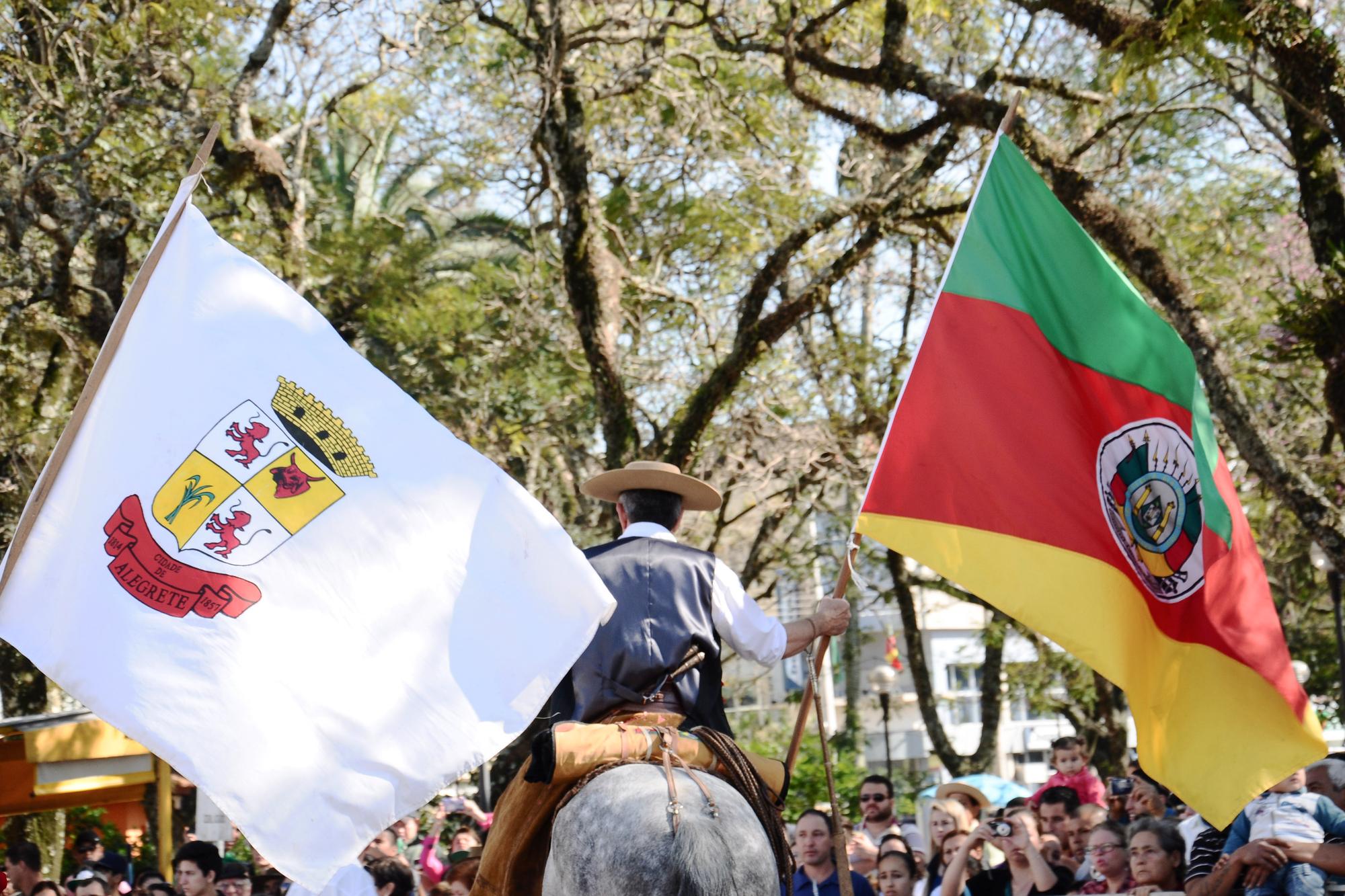 Cavaleiros pilchados abrindo o desfile de 20 de Setembro em Alegrete, um com a bandeira do Rio Grande do Sul e outro com a bandeira da cidade, diante do público na calçada, sob as árvores.