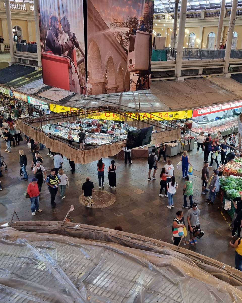 Interior do Mercado Público de Porto Alegre visto do mezanino, com bancas de frutas e peixes, corredores cheios de gente e as arcadas amarelas do prédio.