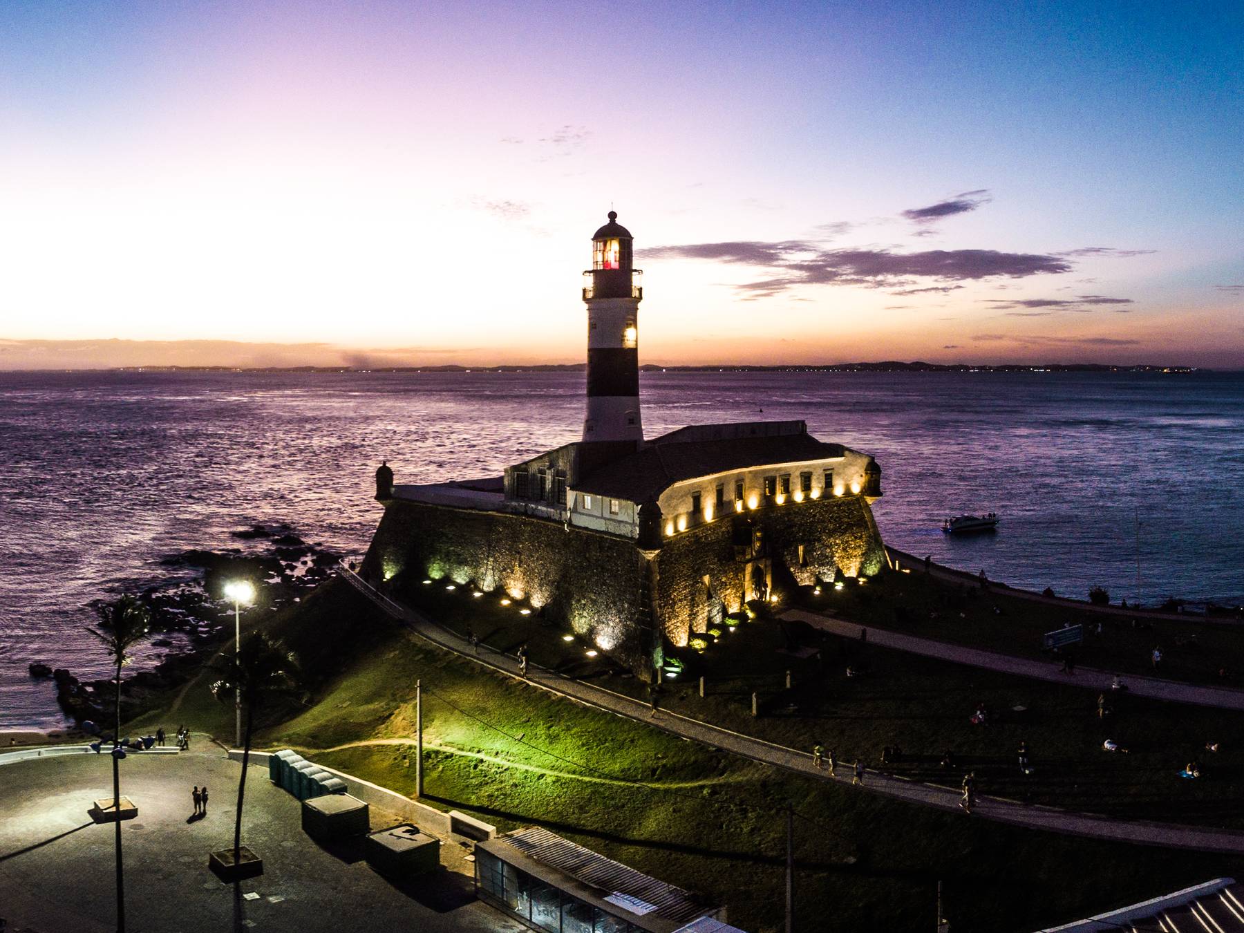 Farol da Barra e o Forte de Santo Antônio iluminados ao anoitecer, em Salvador, vistos do alto, com o céu lilás, o mar da Baía de Todos-os-Santos e o calçadão da orla com postes acesos.