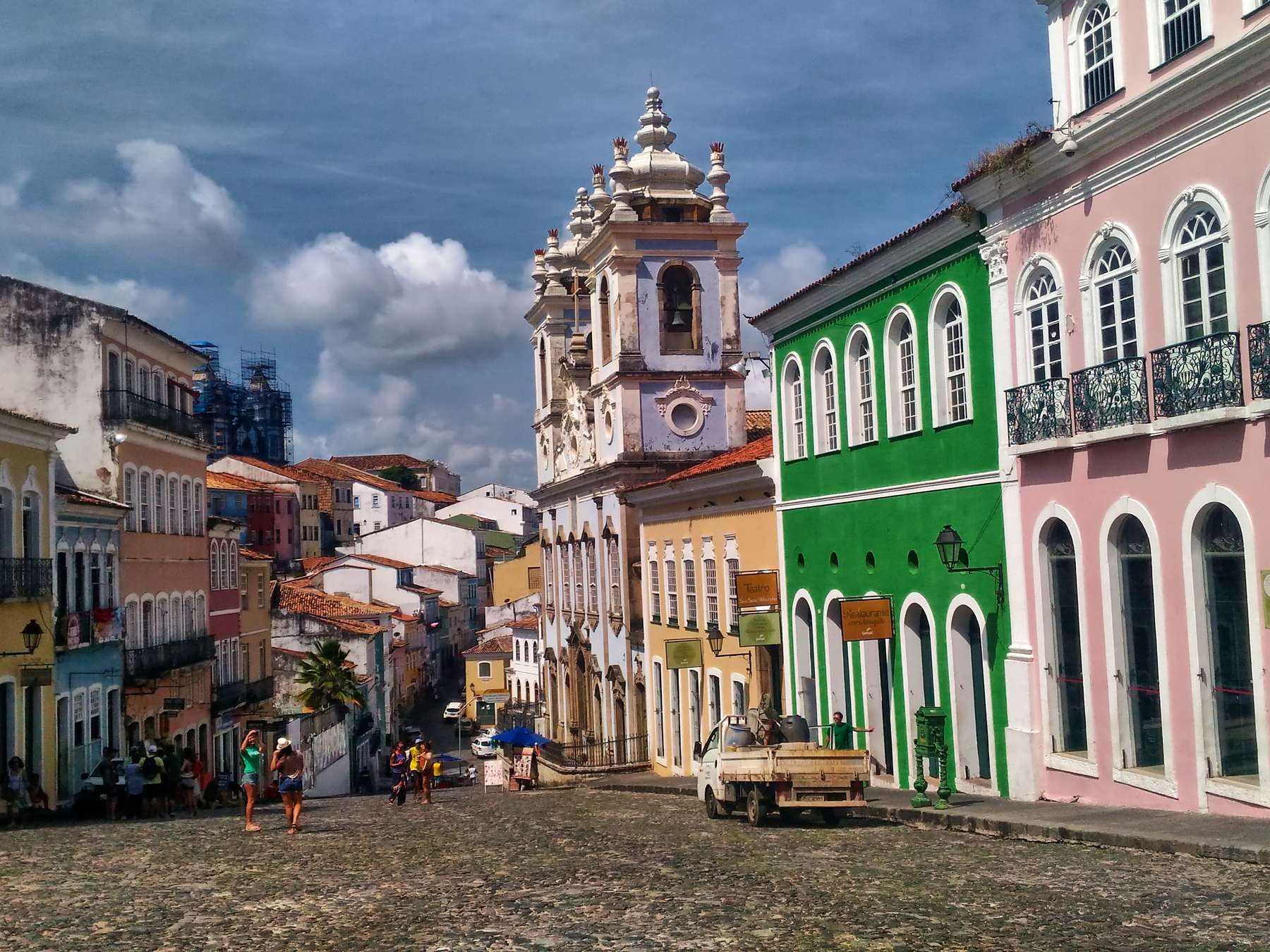 Largo do Pelourinho, em Salvador, com casarões coloniais verdes, rosa e amarelos, a Igreja do Rosário dos Pretos ao fundo, o calçamento de pedra e pessoas caminhando sob um céu de nuvens.