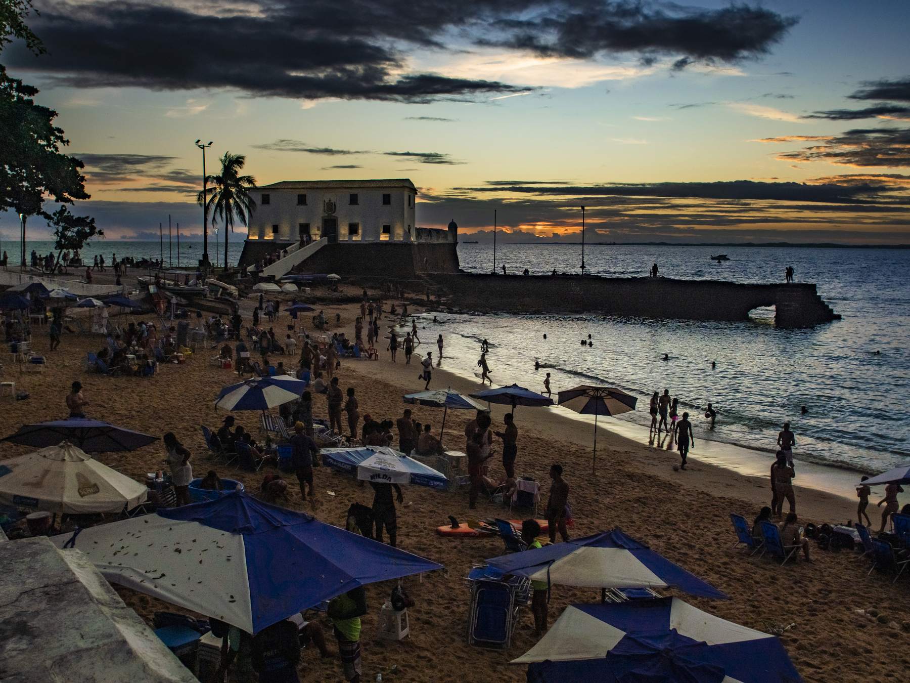 Praia do Porto da Barra, em Salvador, cheia de gente ao pôr do sol, com guarda-sóis azuis na areia, banhistas no mar calmo e o Forte de Santa Maria contra o céu alaranjado.