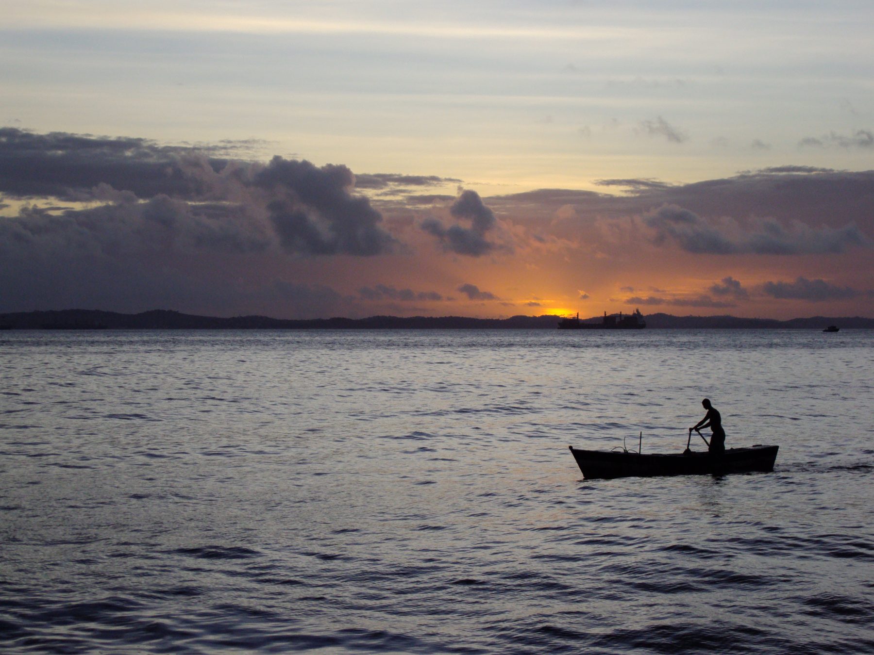 Pescador remando uma canoa em silhueta na Baía de Todos-os-Santos, em frente ao Solar do Unhão, em Salvador, com o sol se pondo entre nuvens escuras no horizonte.
