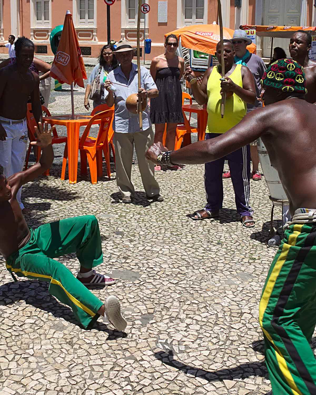Roda de capoeira no calçamento de pedras portuguesas do Terreiro de Jesus, em Salvador, com dois capoeiristas de calça verde e amarela jogando e o público em volta, num dia de sol.