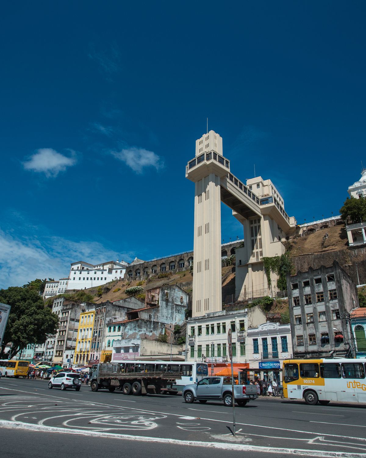 Torre do Elevador Lacerda vista da Praça Cairu, na Cidade Baixa de Salvador, com a encosta da Cidade Alta, os casarões antigos, ônibus e carros na avenida e o céu azul.