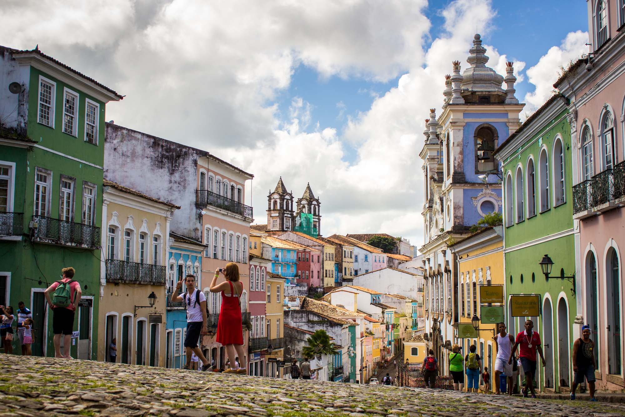 Turistas e moradores caminhando pelo Largo do Pelourinho, em Salvador, entre casarões coloniais verdes, azuis, amarelos e rosa, com a torre azul da Igreja do Rosário dos Pretos e o céu de nuvens brancas ao fundo.