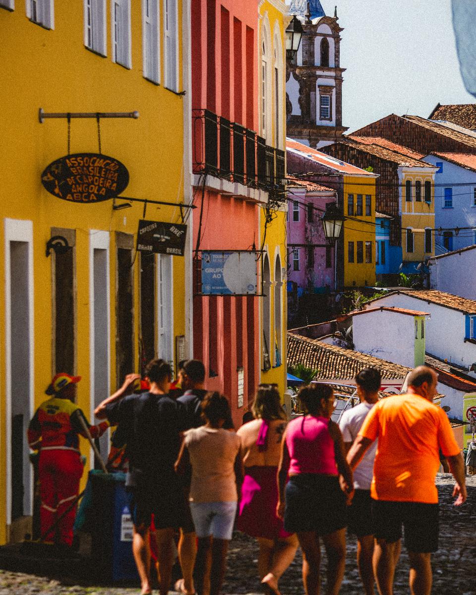Grupo de turistas de bermuda e camiseta subindo a ladeira de pedra do Pelourinho, em Salvador, entre casarões amarelos, laranja e azuis, com a torre de uma igreja ao fundo e um gari de uniforme laranja à esquerda.