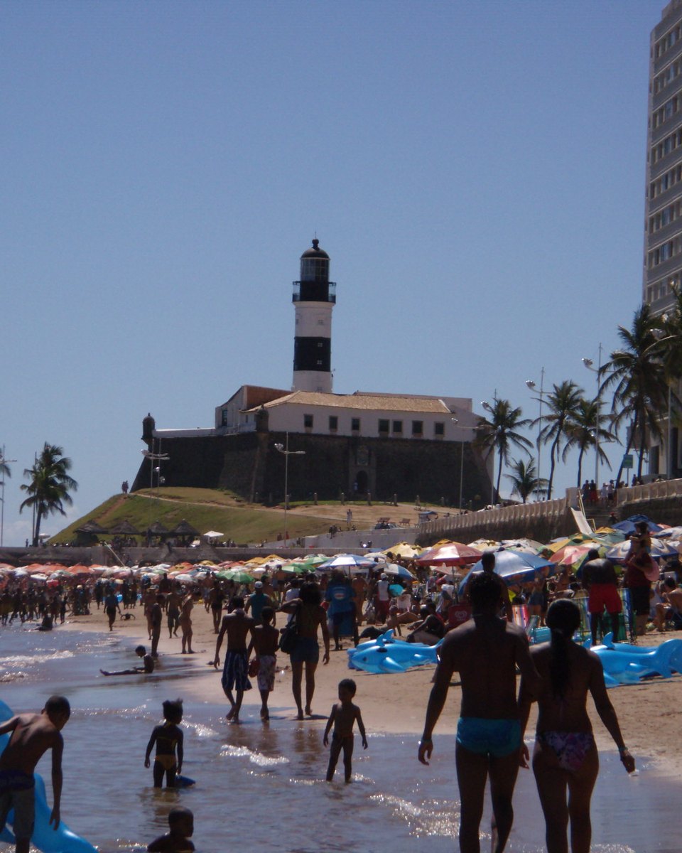Praia do Farol da Barra, em Salvador, cheia de banhistas em um dia de sol, com guarda-sóis coloridos na areia, gente entrando no mar e o farol listrado de preto e branco sobre o forte ao fundo.