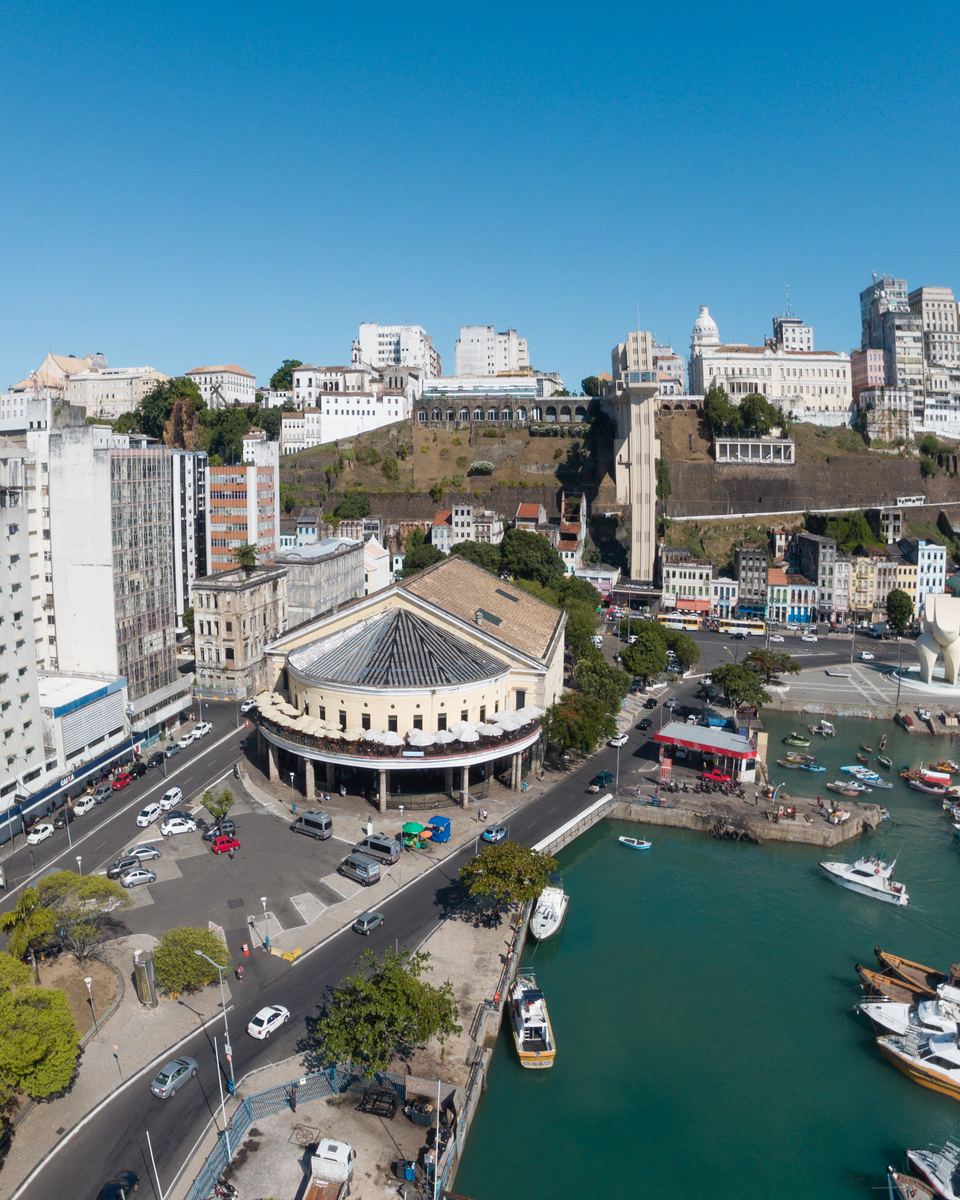 Vista aérea da Cidade Baixa de Salvador, com o prédio arredondado do Mercado Modelo, a marina com barcos, a avenida movimentada e, ao fundo, a encosta verde da Cidade Alta com o Elevador Lacerda e os prédios do Centro.