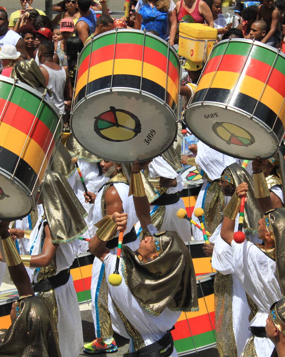 Percussionistas do Olodum com fantasias douradas e brancas erguendo tambores pintados de verde, amarelo, vermelho e preto no meio da multidão, num desfile do Carnaval de Salvador.