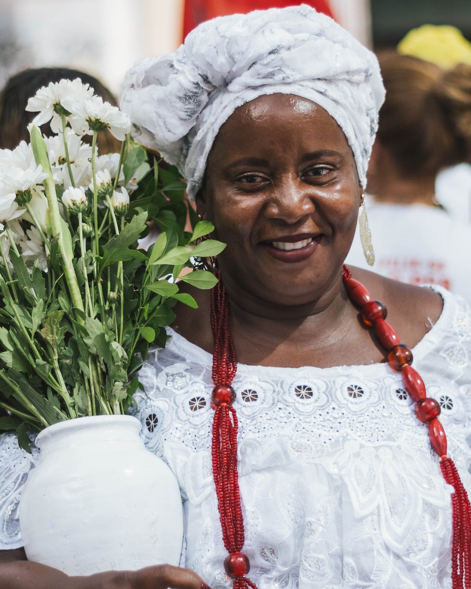 Baiana de turbante branco e colar de contas vermelhas sorrindo com um vaso de flores brancas nas mãos, em Salvador.