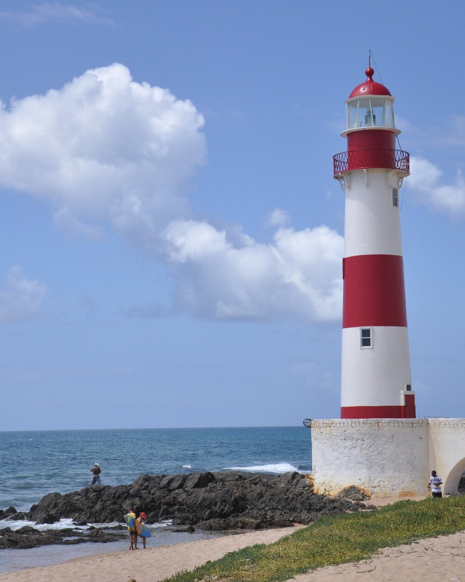 Farol de Itapuã, listrado de vermelho e branco, sobre as pedras da praia em Salvador, com céu azul.
